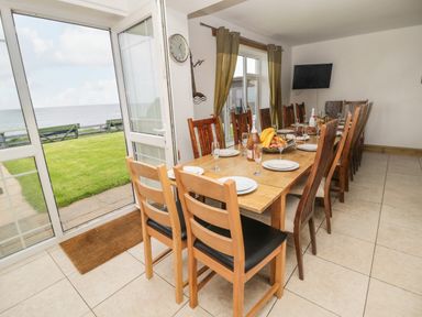 A dining room with a long wooden table set with plates glasses and fruit near glass doors opening to a lawn and sea view at On The Watersedge in Beadnell