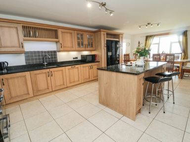 A kitchen with wooden cabinets black countertops a kitchen island with two stools and a dining table with chairs by windows at On The Watersedge in Beadnell