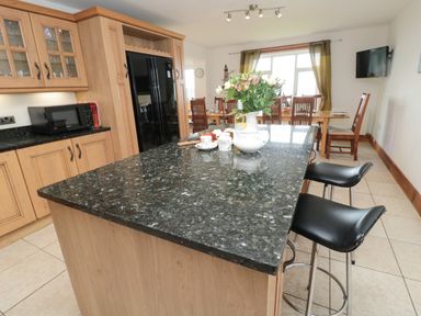 A kitchen with wooden cabinets a black refrigerator black countertop with flowers and cups two black stools and a dining table in the background at On The Watersedge in Beadnell