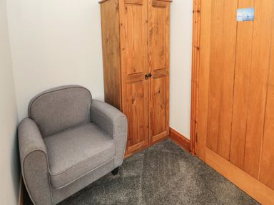 A corner with a gray armchair wooden wardrobe and wooden door on a carpeted floor at On The Watersedge in Beadnell