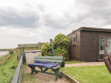A wooden bench with a cushion in a garden next to a brown wooden shed at On The Watersedge in Beadnell