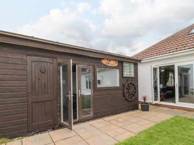 An outdoor patio area with a wooden shack labeled hot tub shack and glass doors opening beside a house with tiled roof at On The Watersedge in Beadnell