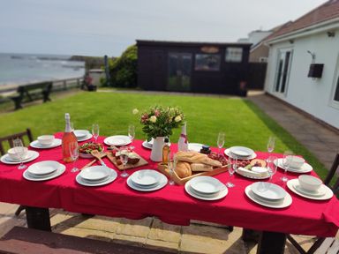 An outdoor dining table with plates glasses bread and flowers on a red tablecloth in a garden near the sea at On The Watersedge in Beadnell