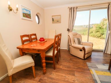 A dining area with a wooden table and chairs next to a beige armchair by a sliding glass door at The Shack in Mawgan Porth near Newquay