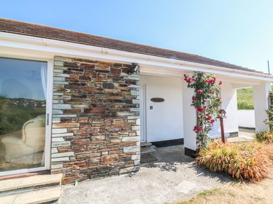 An exterior view of a house with a stone wall and white walls with a covered entryway and climbing rose plants at The Shack in Mawgan Porth near Newquay