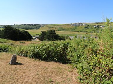 A grassy field with bushes and a stone marker with houses and hills in the background at The Shack in Mawgan Porth near Newquay