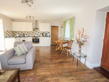 An open plan kitchen and dining area with white cabinets a gray armchair wooden dining table with chairs and a glass side table with a floral arrangement at Middle Barn in Launceston