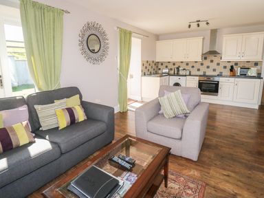 An open living room and kitchen space with a gray sofa and armchair with cushions a glass coffee table and white kitchen cabinets at Middle Barn in Launceston