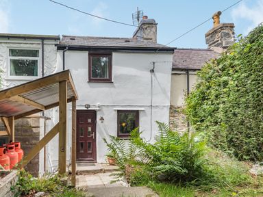 A white two-story house with a brown door and windows a small garden with plants and a covered area with red gas canisters at 3 Tyn Y Mynydd in Penmachno