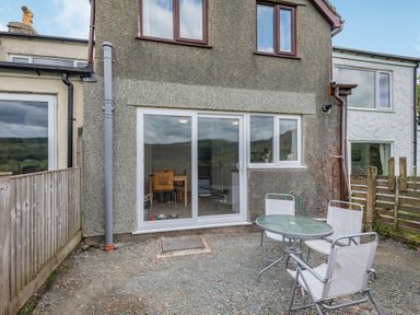 A backyard with a gravel ground a round glass table with four white chairs outside a house with sliding glass doors and two upstairs windows at 3 Tyn Y Mynydd in Penmachno