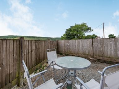 An outdoor patio area with a round glass table and three white chairs surrounded by wooden fencing at 3 Tyn Y Mynydd in Penmachno