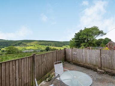 An outdoor patio area with a glass table and chairs next to a wooden fence overlooking green hills and trees at 3 Tyn Y Mynydd in Penmachno
