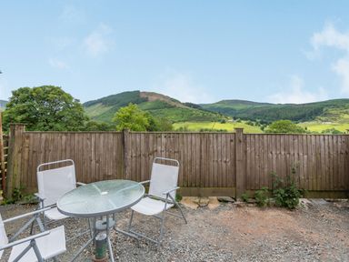 An outdoor patio area with a glass table and three white chairs next to a wooden fence with hills in the background at 3 Tyn Y Mynydd in Penmachno