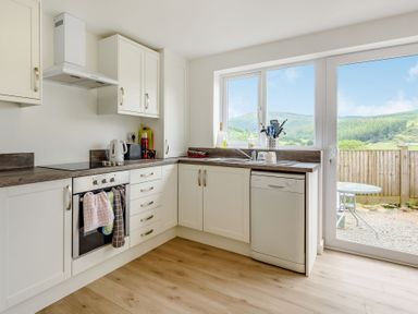 A kitchen with white cabinets a stove a kettle a dishwasher and large windows showing an outdoor table and hills at 3 Tyn Y Mynydd in Penmachno