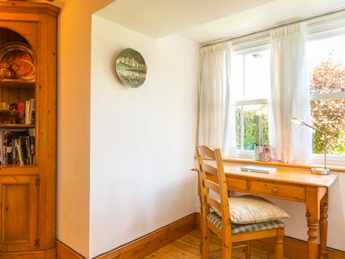 A wooden desk and chair with cushions by a window with white curtains and a wooden bookshelf in a room at Townfoot Cottage in Elsdon near Otterburn