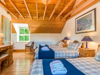 A bedroom with three single beds each with folded towels bedside tables with lamps a wicker chair and wooden ceiling at Townfoot Cottage in Elsdon near Otterburn