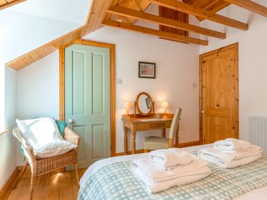 A bedroom with a bed topped with folded towels a wooden dressing table with lamps and a wicker chair near a light green door at Townfoot Cottage in Elsdon near Otterburn