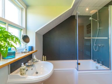 A bathroom with a bathtub and shower combination a sink with two faucets and a window sill with a plant mirror and soap bottles at Townfoot Cottage Elsdon near Otterburn