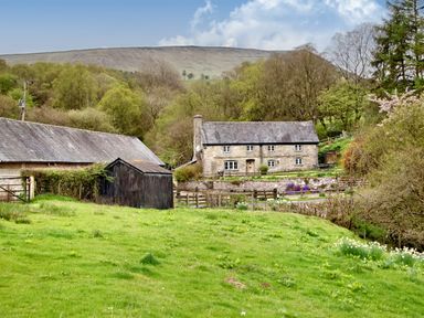 A stone house with adjacent barns on a grassy hillside surrounded by trees and hills at The Birches in Hay-on-Wye