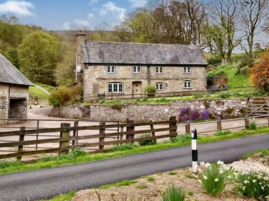 A stone cottage with white windows and a slate roof behind a wooden fence and stone wall at The Birches in Hay-on-Wye