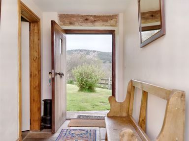 An open wooden front door showing a grassy yard with a bush and hills beyond a hallway with a wooden bench a mirror and rugs at The Birches in Hay-on-Wye