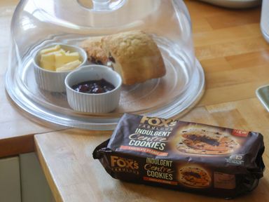 A wooden countertop with a glass dome covering scones and small dishes of butter and jam and a packet of Fox's Indulgent Centre Cookies chocolate orange flavor