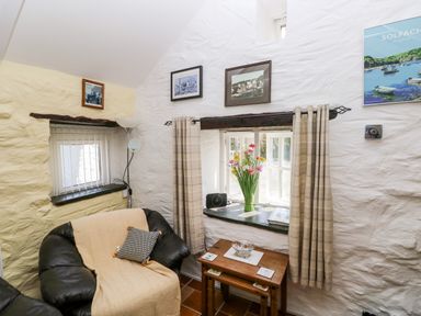 A sitting area with a black leather armchair covered by a yellow throw a small wooden table with a glass bowl flowers in a vase on the windowsill and framed pictures on white stone walls at Ffynnon Tom in Trefin near St Davids