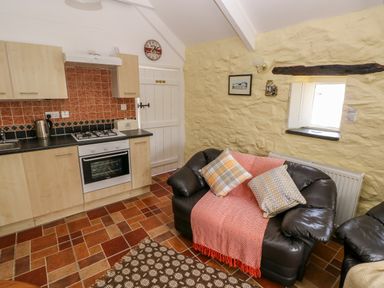 A kitchen and living area with wooden cabinets tiled walls and floor a black leather armchair with cushions and a throw at Ffynnon Tom in Trefin near St Davids