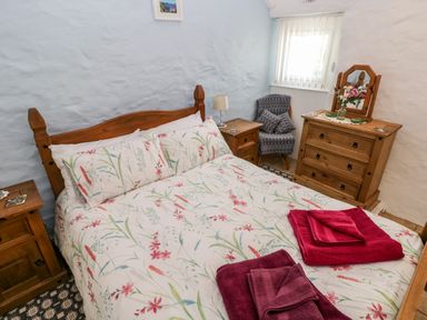 A bedroom with a wooden bed with floral bedding two wooden nightstands a wooden dresser a patterned armchair and folded towels on the bed at Ffynnon Tom in Trefin near St Davids