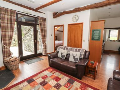 A living room with a leather sofa and plaid pillows next to wooden doors and large glass doors with plaid curtains at The Hayloft in Newlands near Braithwaite and Keswick