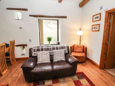 A sitting area with a leather sofa with two plaid cushions a brown round chair a floor lamp and a window with a vase of flowers at The Hayloft in Newlands near Braithwaite and Keswick