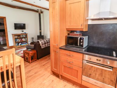 A kitchen with wooden cabinets a microwave and an oven leading into a living area with a sofa a wood stove and a bookcase at The Hayloft in Newlands near Braithwaite and Keswick