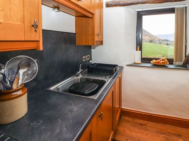 A kitchen counter with a sink drying rack and utensils in a pot next to a window showing a grassy hillside at The Hayloft in Newlands near Braithwaite and Keswick