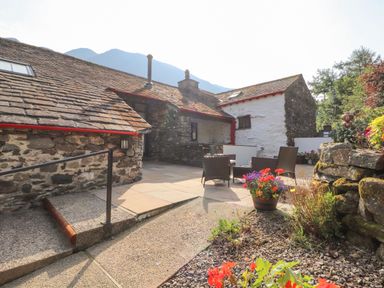An outdoor patio area with stone walls and slate roof a seating area with chairs and a table flower pots with colorful flowers and a stone pathway at The Hayloft in Newlands near Braithwaite and Keswick