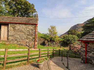 A stone building with a wooden door next to a wooden fence and a potted flower under a blue sky at The Hayloft in Newlands near Braithwaite and Keswick