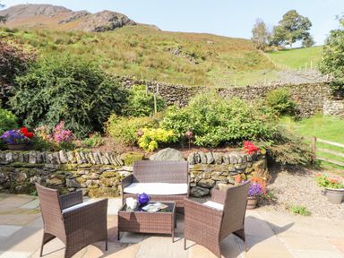 A patio with wicker chairs and a table with a teapot and cups in a garden with stone walls and plants at The Hayloft in Newlands near Braithwaite and Keswick