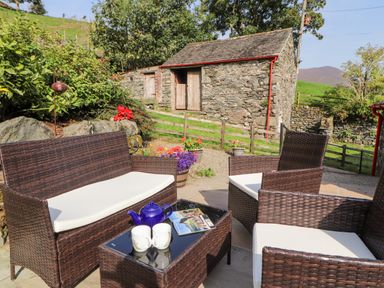 An outdoor seating area with brown wicker furniture and white cushions a blue teapot and two white mugs on a glass table stone building and green hill in the background at The Hayloft in Newlands near Braithwaite and Keswick