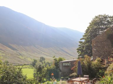 An outdoor patio with a table and chairs and a closed umbrella next to a stone building overlooking hills and greenery at The Hayloft in Newlands near Braithwaite and Keswick