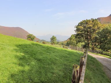 A grassy field with a wooden fence and a dirt path with trees and hills in the background at The Hayloft in Newlands near Braithwaite and Keswick
