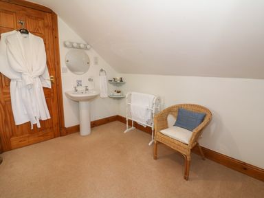 A bathroom with a wooden door a white bathrobe hanging on the door a pedestal sink a round mirror above the sink a wicker chair with cushions a towel rack with white towels and corner shelves holding toiletries at Tangaer Cottage in Cellan near Lampeter