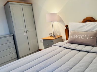 A bedroom with a bed with a wooden headboard a pillow with cwtch on it a bedside table with a lamp and an alarm clock and grey wardrobe and chest of drawers at Tangaer Cottage in Cellan near Lampeter