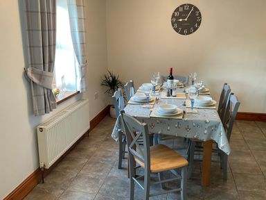 A dining room with a table set for six near a window with curtains and a wall clock at Tangaer Cottage in Cellan near Lampeter