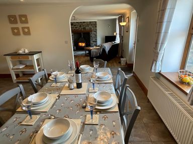 A dining table set with plates glasses and a bottle of wine near a living room with a wood stove at Tangaer Cottage in Cellan near Lampeter
