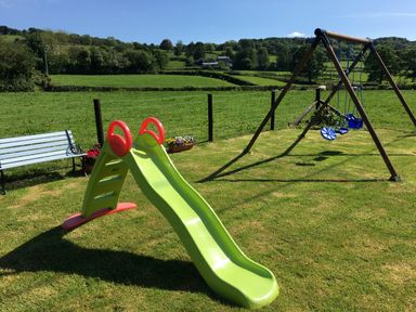 A green slide and a wooden swing set on a grassy lawn with a bench and flowers at Tangaer Cottage in Cellan near Lampeter