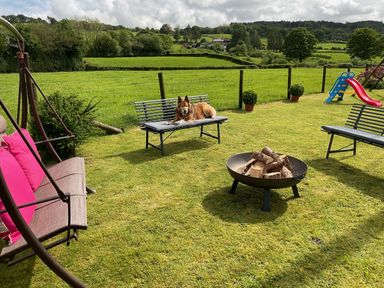 A garden with lawn chairs a wood fire pit a dog lying on a bench and a children's slide at Tangaer Cottage Cellan near Lampeter