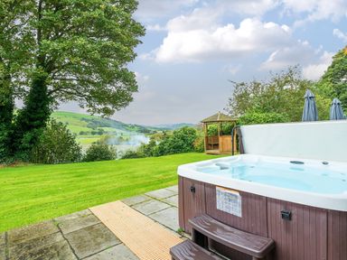 An outdoor hot tub on a stone patio next to a grassy lawn with a wooden gazebo and hills in the background at Old Rectory Cottages Aberhafesp near Newtown
