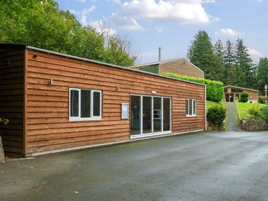 A wooden building with windows and glass doors by a paved area surrounded by trees and other wooden structures at Old Rectory Cottages Aberhafesp near Newtown