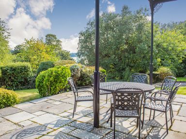 A patio with a round metal table and six metal chairs surrounded by bushes and trees at Menai View in Llanfairpwllgwyngyll near Beaumaris