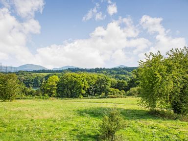 A grassy field with trees and hills in the background under a partly cloudy sky at Menai View in Llanfairpwllgwyngyll near Beaumaris