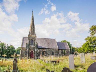 A stone church with a tall steeple in a graveyard with headstones and trees